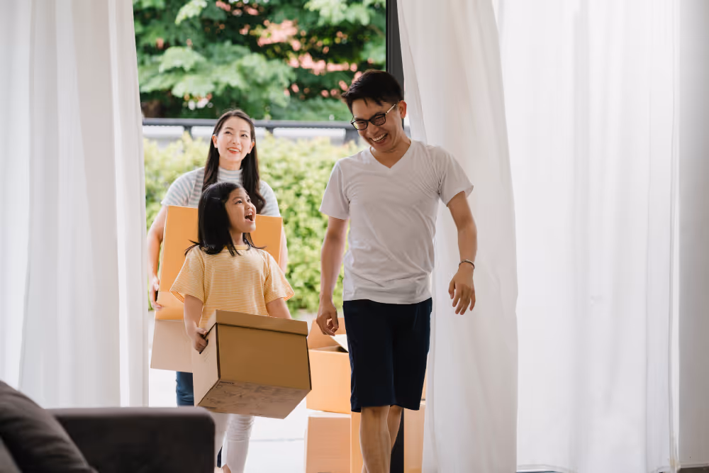 A family of three happily moves into a new home, carrying boxes with trees and sunlight behind them.
